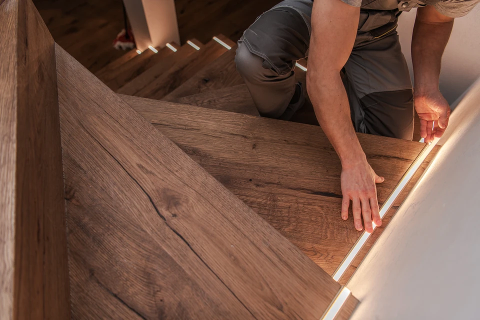 Photographie d'un artisan menuisier expérimenté en discussion avec un client dans un atelier de menuiserie traditionnelle. L'artisan, vêtu de manière professionnelle (gilet ou tablier de travail), montre des plans ou esquisses d'escalier sur une table de travail entourée d'outils et de bois. L'atmosphère doit être chaleureuse et professionnelle, reflétant l'engagement, l'écoute et l'expertise artisanale. L'environnement doit montrer des éléments de bois travaillé, des machines traditionnelles ou des pièces de menuiserie en arrière-plan pour authentifier le savoir-faire artisanal bois.