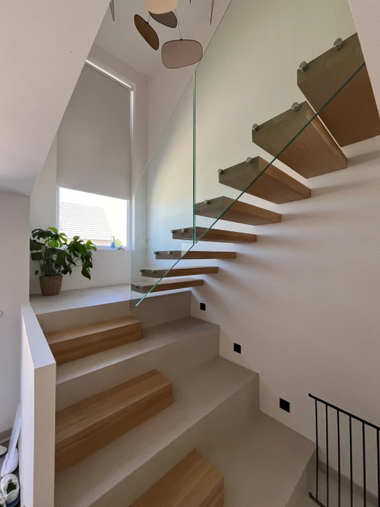 Vue d'un intérieur moderne et épuré montrant un escalier suspendu contemporain en bois clair et métal noir, avec marches flottantes et rampe minimale, photographié dans un loft ou maison contemporaine avec grande hauteur sous plafond. L'escalier doit être au centre de la composition, intégré harmonieusement dans un espace lumineux avec murs clairs, créant une sensation de légèreté et d'espace aéré. La photographie doit capturer l'élégance du design et la transformation de l'espace intérieur.